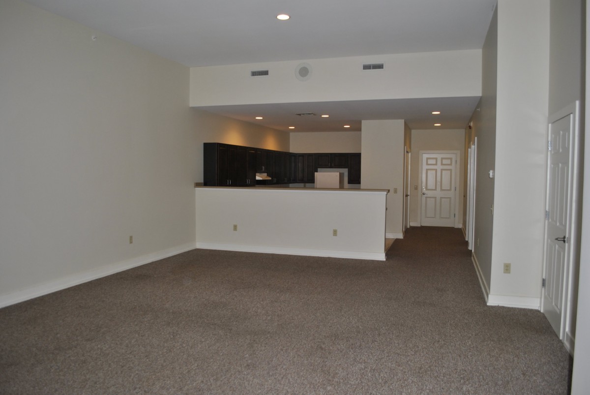 An empty apartment living room with a view of the kitchen area, featuring carpeted flooring and recessed lighting.