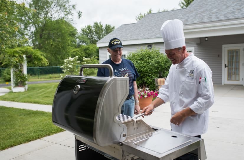 A chef in a white uniform prepares food on a grill while another person observes in a backyard setting.