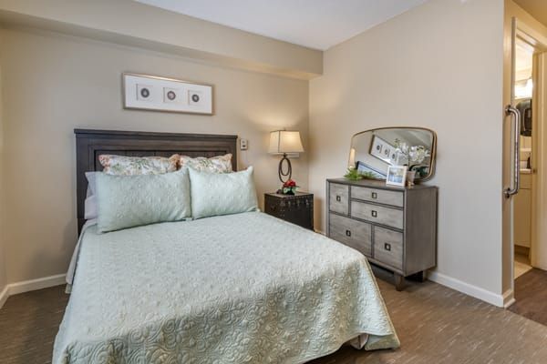 A cozy bedroom featuring a neatly made bed with patterned bedding, a wooden dresser, and decorative elements such as a lamp and wall art.