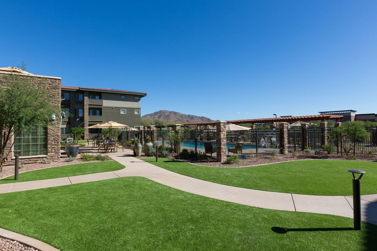 A well-maintained outdoor area with a modern residential building, featuring a swimming pool, palm trees, and green lawns under a clear blue sky.