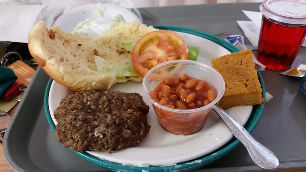 A plate containing a burger patty, lettuce, tomato slices, baked beans in a cup, and cornbread.
