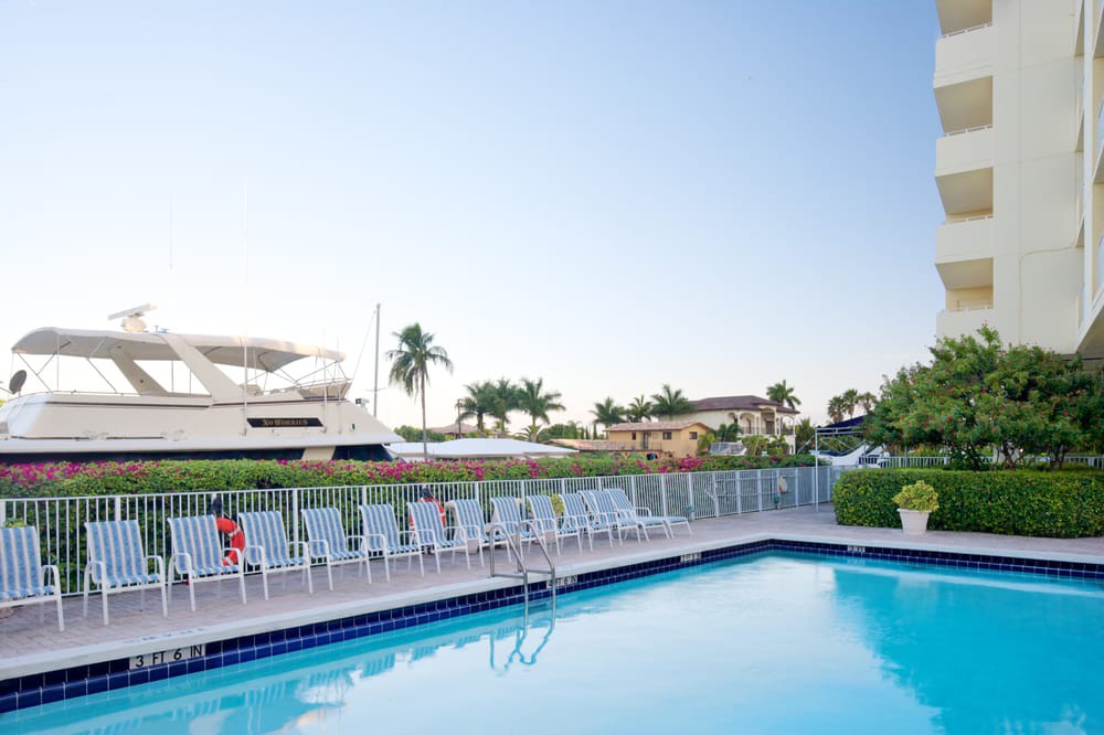 A serene outdoor pool area with lounge chairs, a yacht docked nearby, and palm trees under a clear sky.