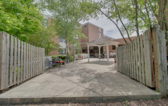 A peaceful courtyard featuring trees and a paved walkway leading to a building with an entrance covered by a canopy.
