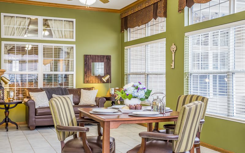 A cozy dining area with green walls, featuring striped chairs and a well-set table under natural light from large windows.