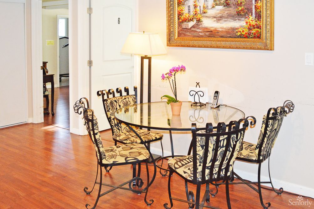 A cozy dining area with a glass-top table and ornate chairs, decorated with floral cushion seats, sits on a hardwood floor beneath elegant artwork. A lamp and an orchid plant add warmth to the space.