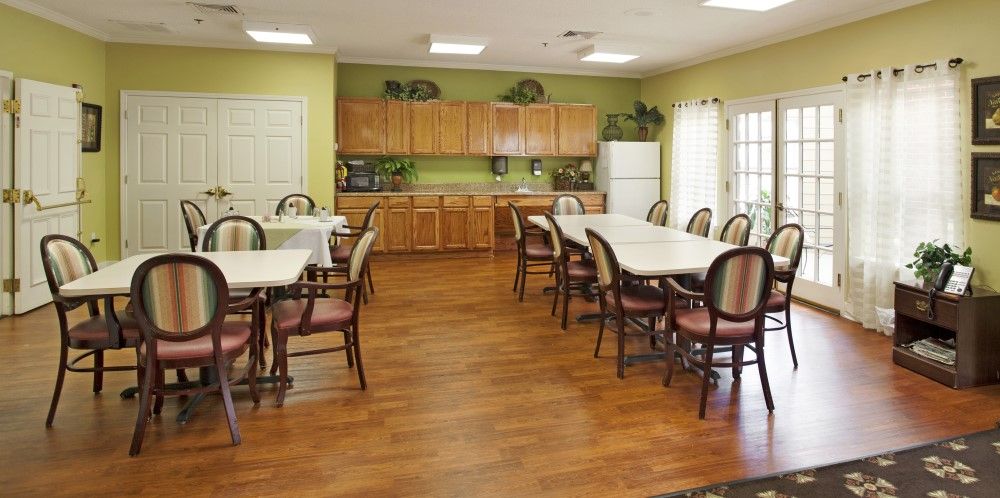 A spacious dining area with multiple tables and chairs, featuring wooden flooring and a kitchen setup with cabinets and a refrigerator.