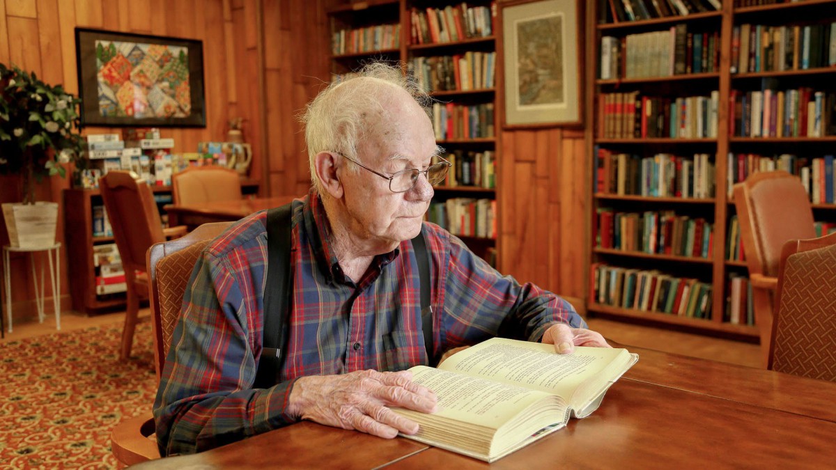 An elderly man wearing glasses and suspenders reads a book in a cozy library setting.