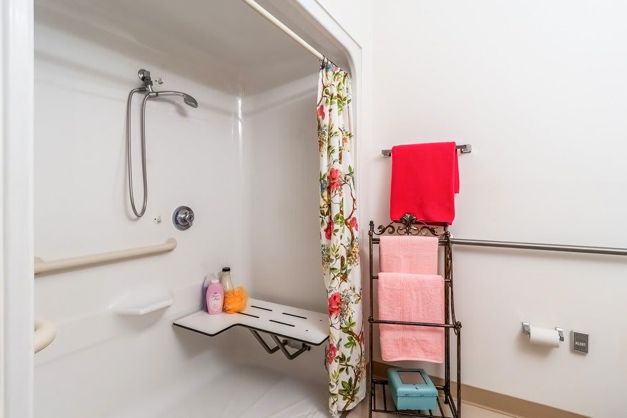 An accessible bathroom with a foldable seat and handheld shower head, featuring a floral shower curtain and colorful towel rack.