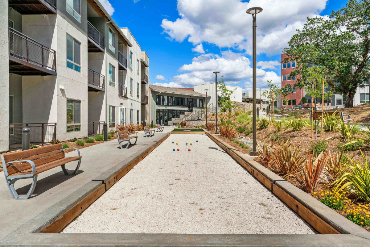 A modern apartment complex features an outdoor bocce court surrounded by benches and landscaping under a bright blue sky.