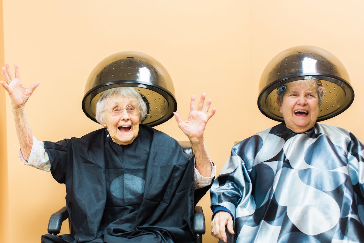 Two elderly women laughing under hair dryers at a salon.