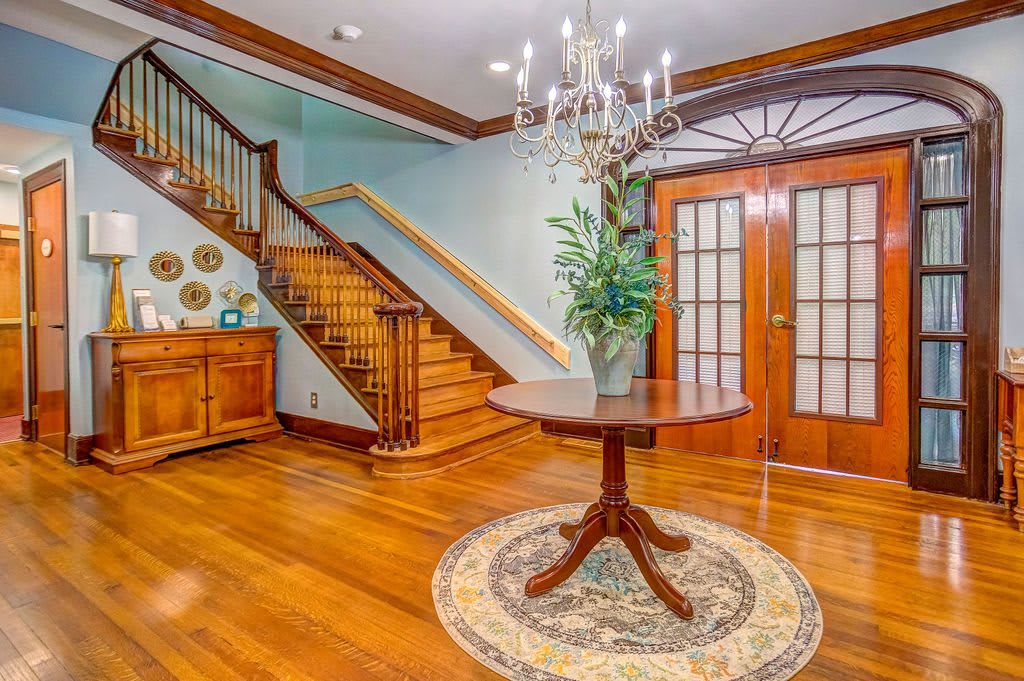 A welcoming foyer with a hardwood floor, featuring a central round table adorned with an indoor plant, under a decorative chandelier. Stairs lead upwards, and French doors offer additional access.