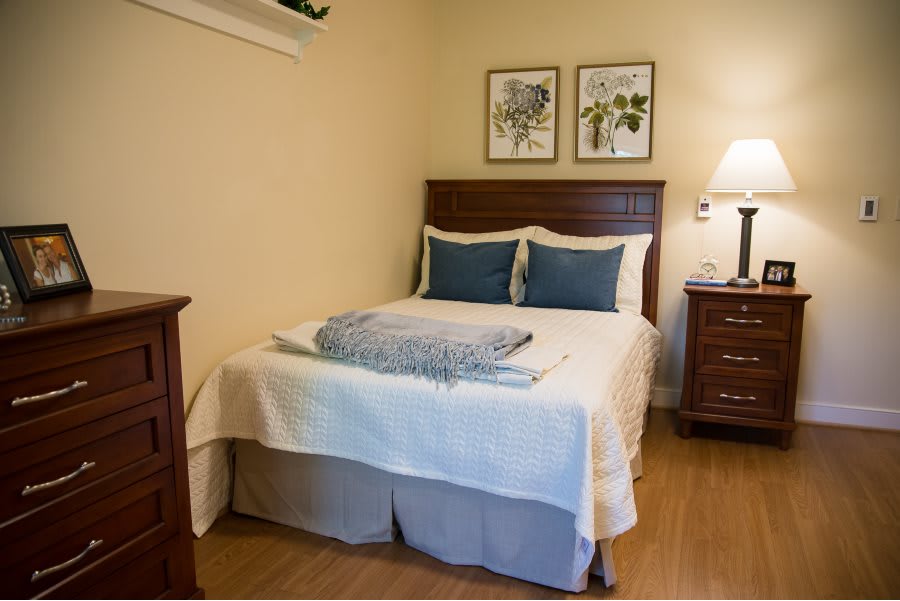 A cozy bedroom featuring a neatly made bed with two blue pillows, a blanket, wooden furniture, and framed artwork on the wall.