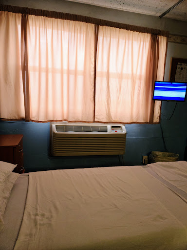 A cozy bedroom featuring a neatly made bed, a window with soft curtains, an air conditioner, and a television.