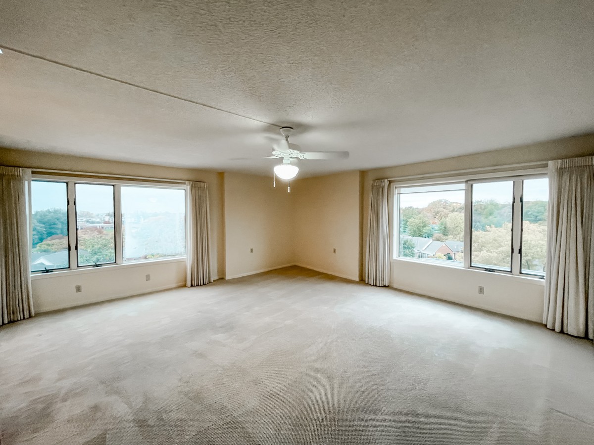 An empty room with large windows, carpet flooring, and a ceiling fan.