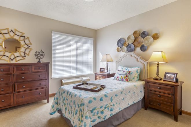 A cozy bedroom with a single bed featuring floral bedding and decorative pillows, flanked by wooden dressers and a lamp. A window with blinds allows natural light to fill the room, complemented by unique wall decor above the headboard.