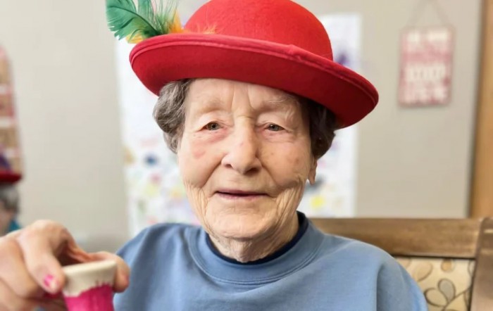 An elderly woman wearing a red hat with a feather, dressed in a blue sweater, smiles for the camera.