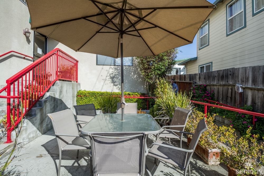 A sunny backyard patio with a table, chairs, and an umbrella surrounded by plants.