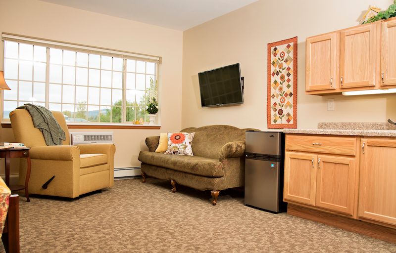 A cozy living room with a couch, armchair, and a mounted television, complemented by wall art and a mini fridge.