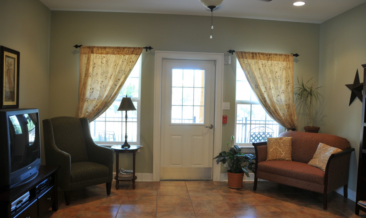 A cozy living room with a central door, flanked by windows adorned with curtains, and featuring comfortable seating and decor.