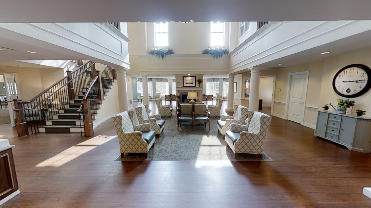 A spacious living room with elegant furniture, featuring multiple chairs and sofas arranged around a central area rug, with large windows allowing natural light to fill the space.