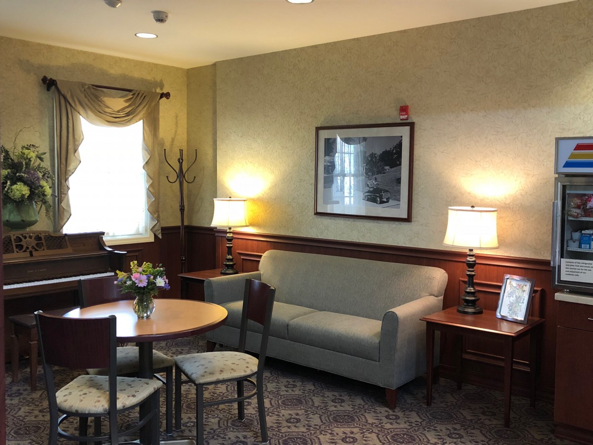 A cozy living room with a sofa, table, chairs, and lamps, featuring artwork on the wall and a window with drapes.