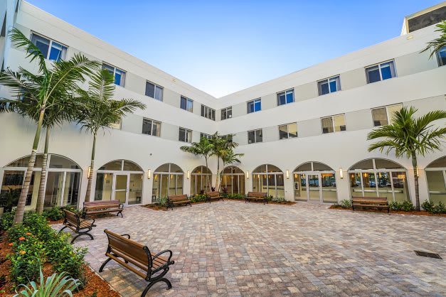 A serene courtyard featuring benches, palm trees, and a building with arched windows.