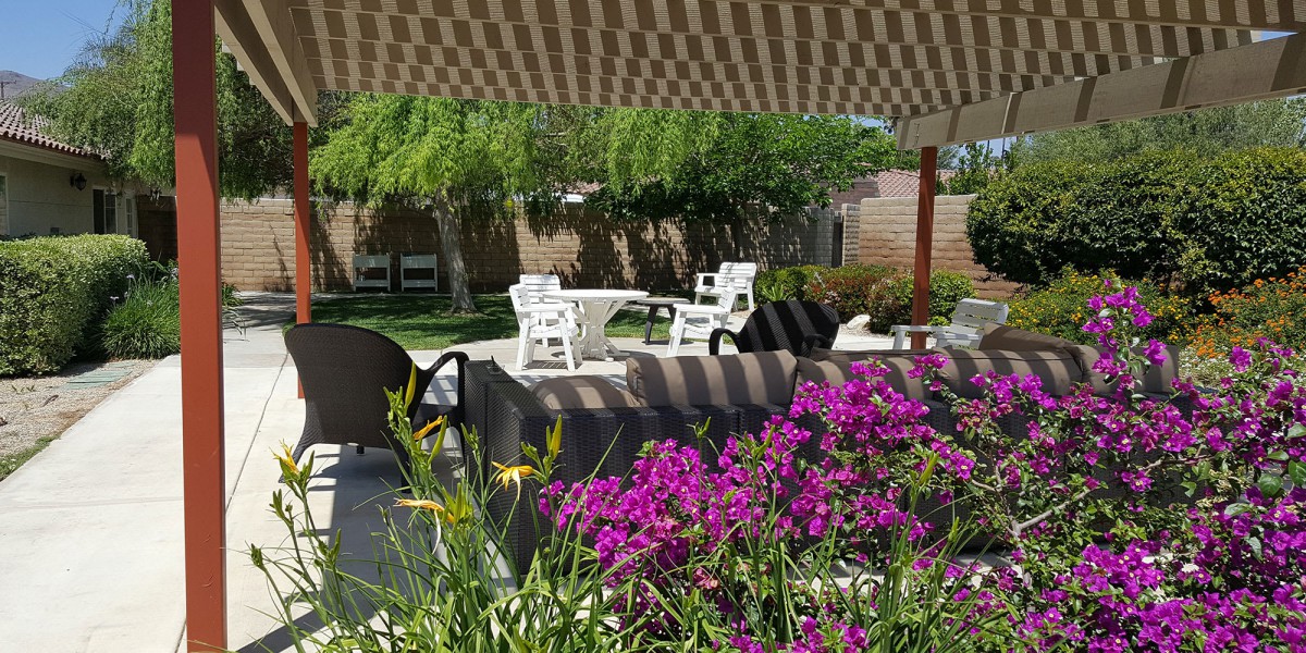 A serene garden setting with outdoor furniture under a shade canopy, surrounded by vibrant flowers and greenery.