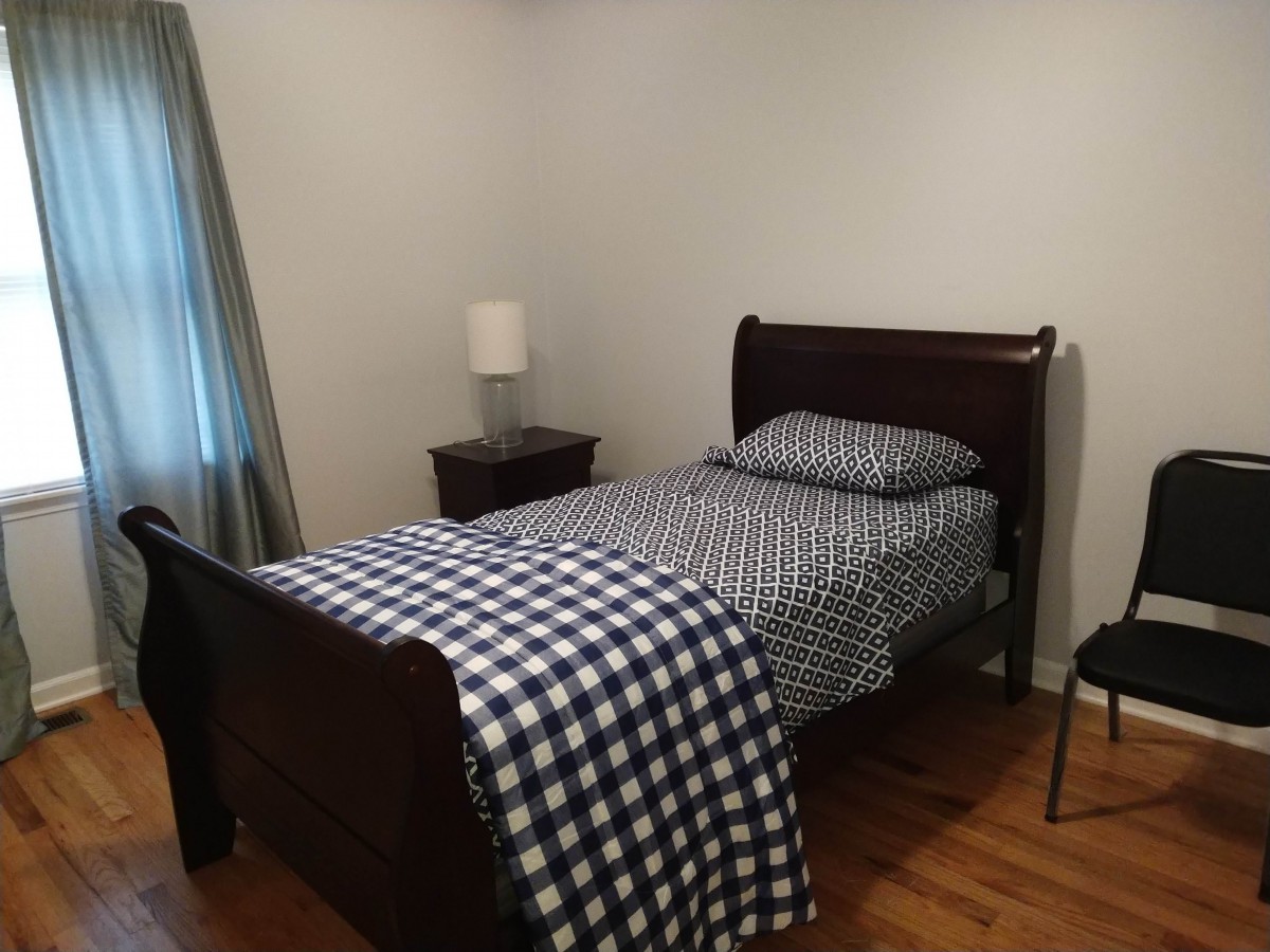 A simple bedroom with a single bed featuring checkered bedding, a nightstand with a lamp, and hardwood flooring.