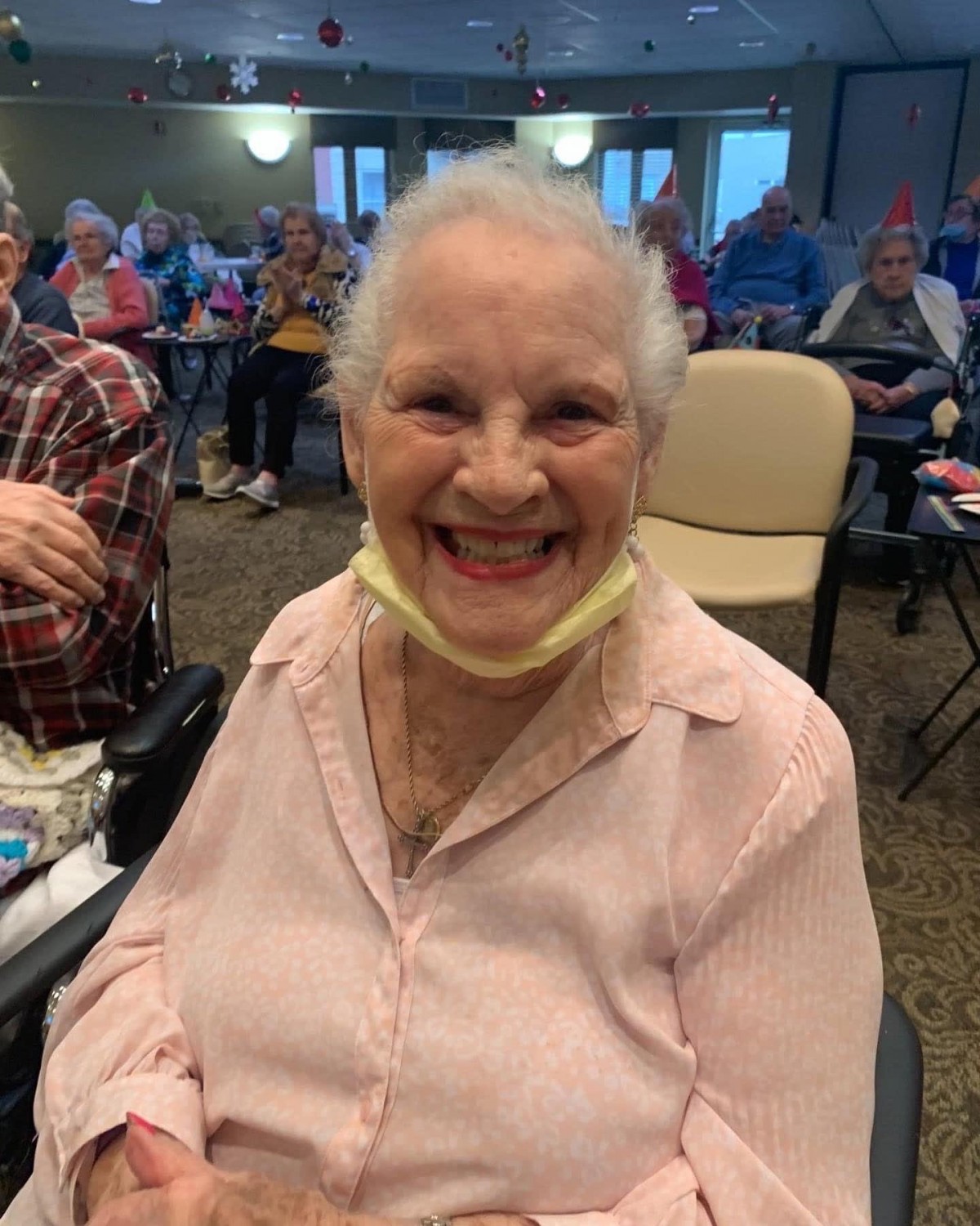 An elderly woman smiling joyfully indoors with a mask resting under her chin, surrounded by an audience.