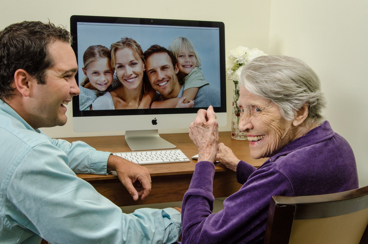 An elderly woman smiles at a family photo displayed on a computer screen, highlighting the use of technology to connect with loved ones.