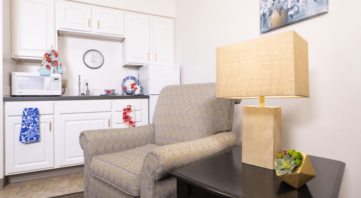 A cozy living room with an armchair and a lamp beside a small kitchenette.