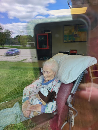An elderly woman sitting in a chair, with a teddy bear, viewed through a reflective window.
