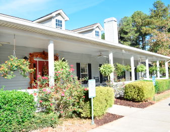 A bright and sunny view of a house with a spacious porch, surrounded by landscaped greenery.