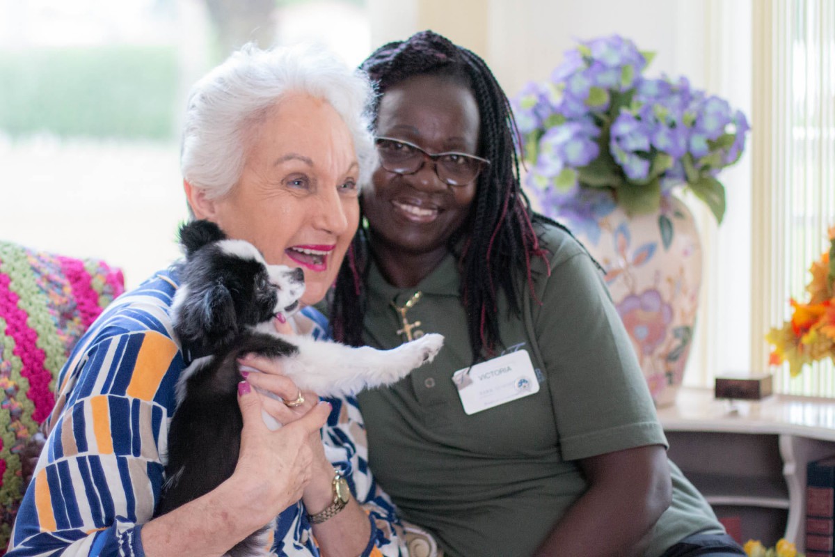 An elderly woman is joyfully holding a small dog while sitting next to her caregiver, both smiling warmly.