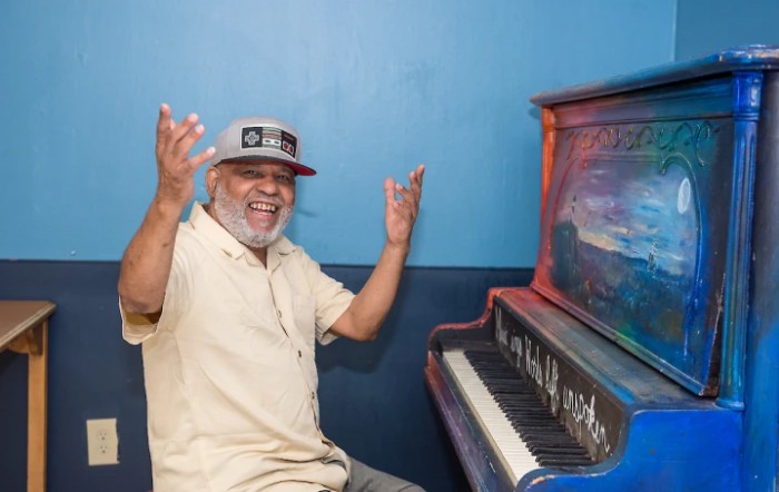 A joyful man wearing a hat sits beside a painted piano, raising his arms in excitement.