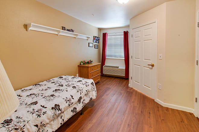 A cozy bedroom with a single bed, dresser, and window adorned with red curtains.