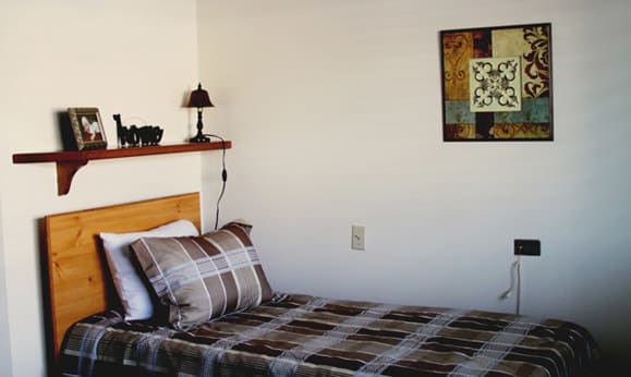 A cozy bedroom with a plaid-patterned bedspread, a decorative shelf with a lamp, and wall art.