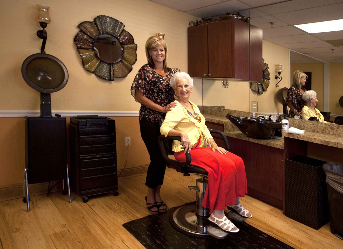 An elderly woman sits in a salon chair, smiling as a hairdresser stands behind her.