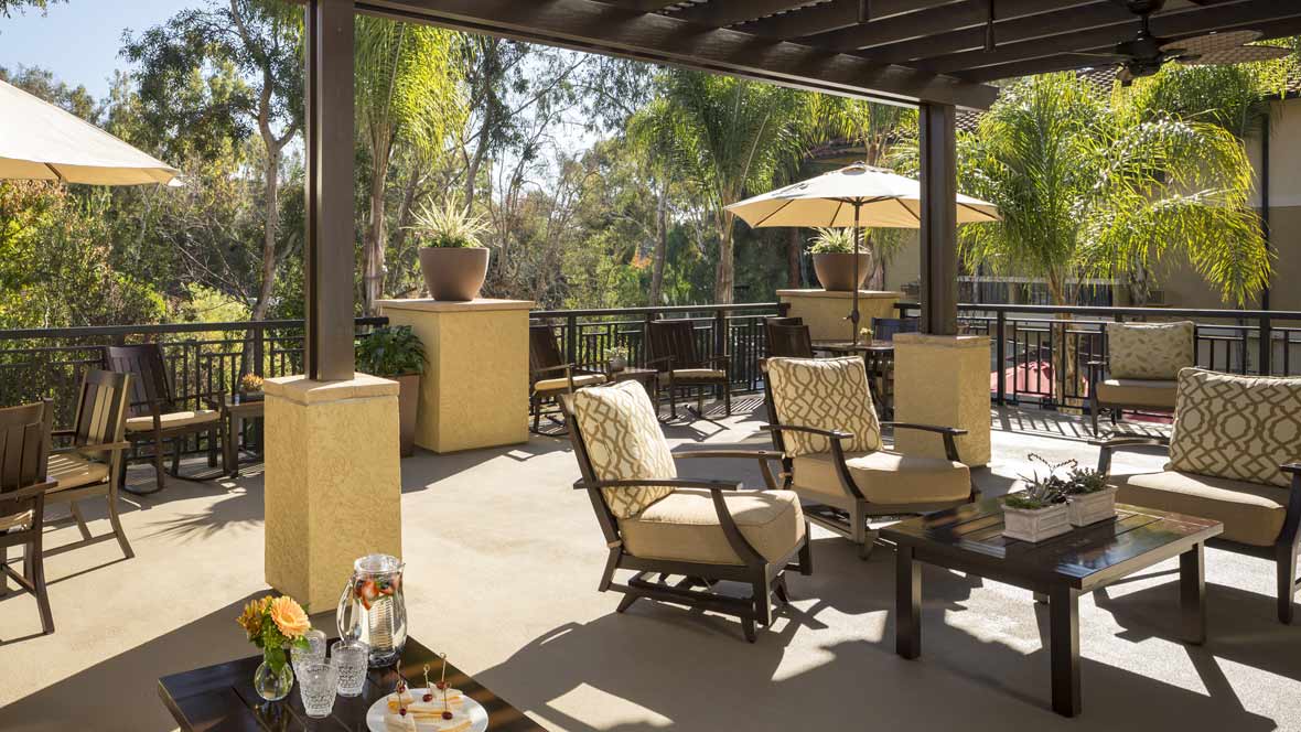 A sunny outdoor patio with comfortable seating and an umbrella, surrounded by lush greenery.