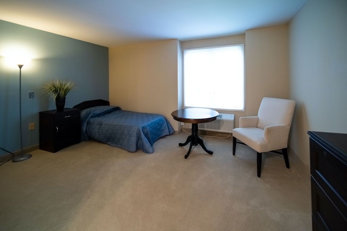 A minimalist bedroom features a bed, chair, and round table with a large window providing natural light.