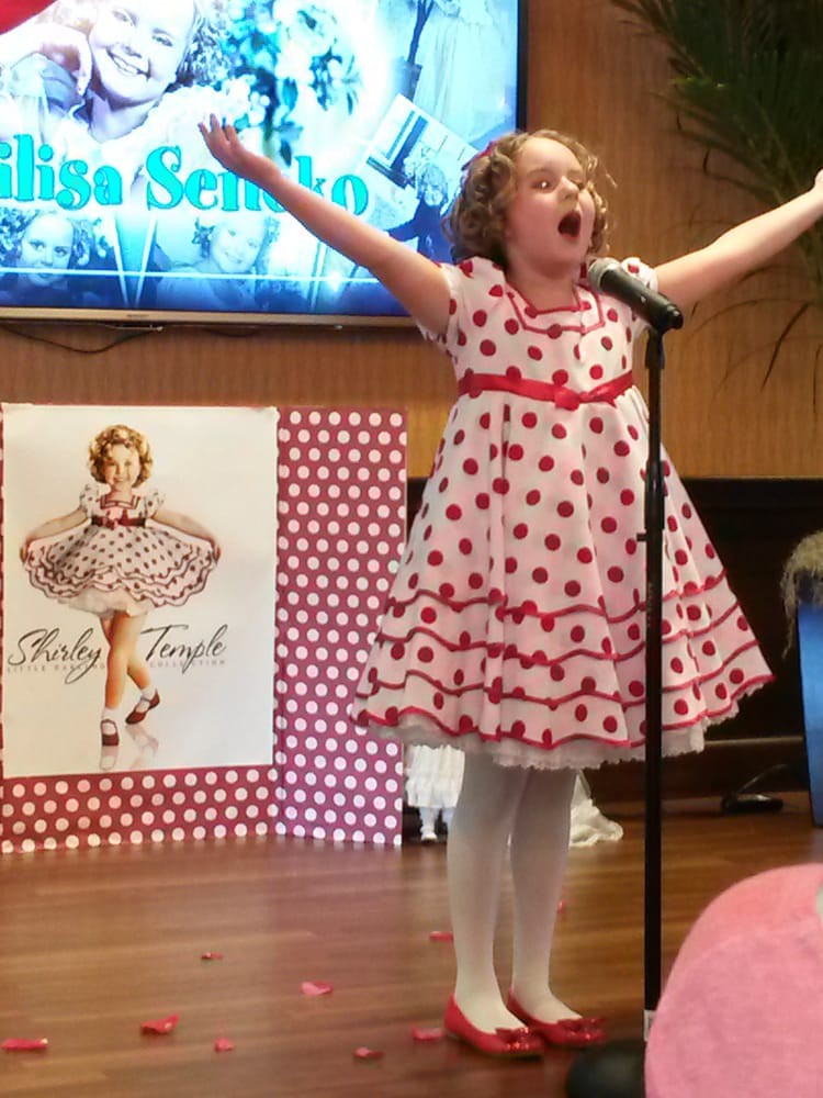 A young girl enthusiastically sings on stage, dressed in a polka dot dress, during an event.