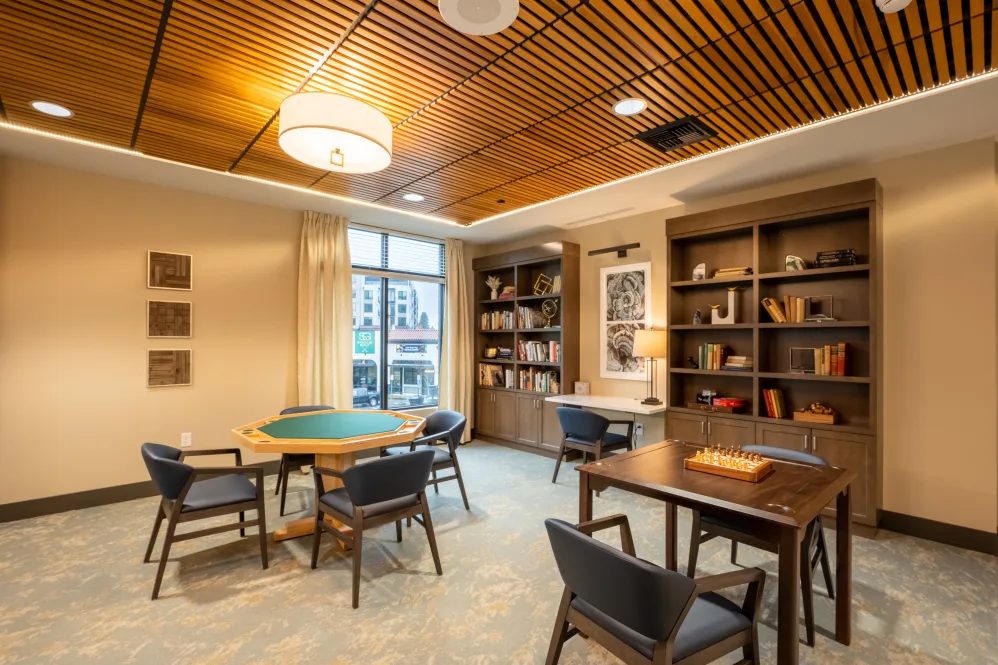 A cozy study room with bookshelves, tables, and chairs, featuring a warm wooden ceiling and soft lighting.