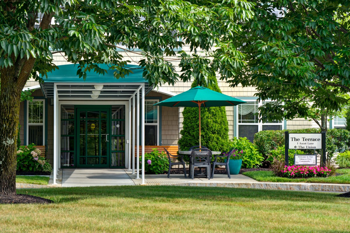 A cozy outdoor seating area with a green umbrella and canopy, surrounded by lush greenery in front of a building.