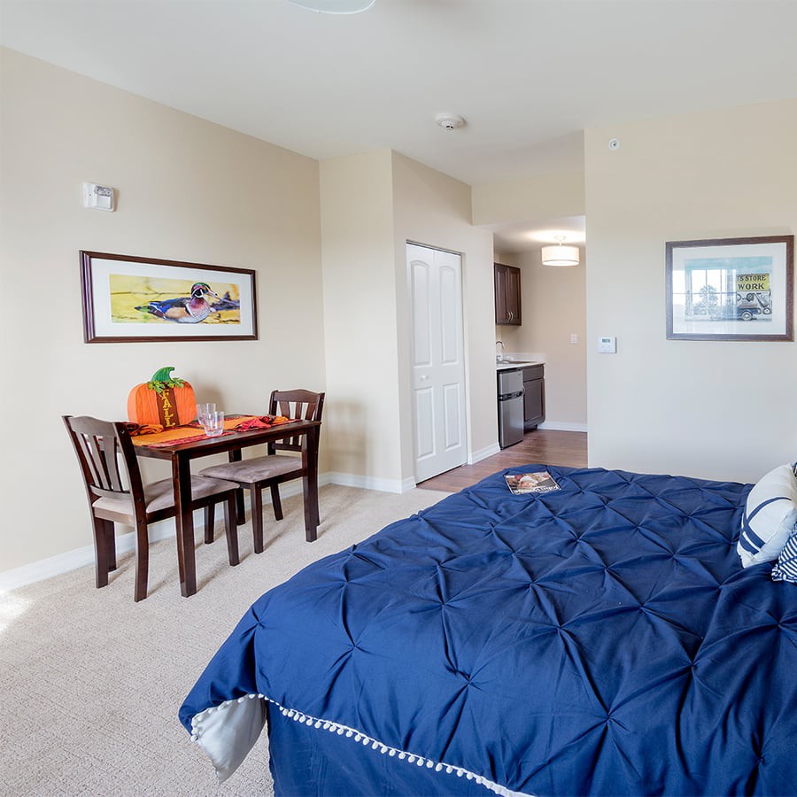 A cozy bedroom featuring a bed with a blue comforter, a small dining table with chairs, and framed artwork on the walls.