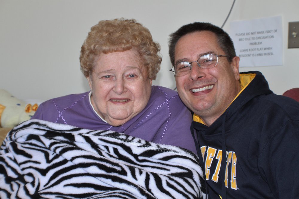 A smiling elderly woman and middle-aged man pose together, holding a zebra-striped blanket.