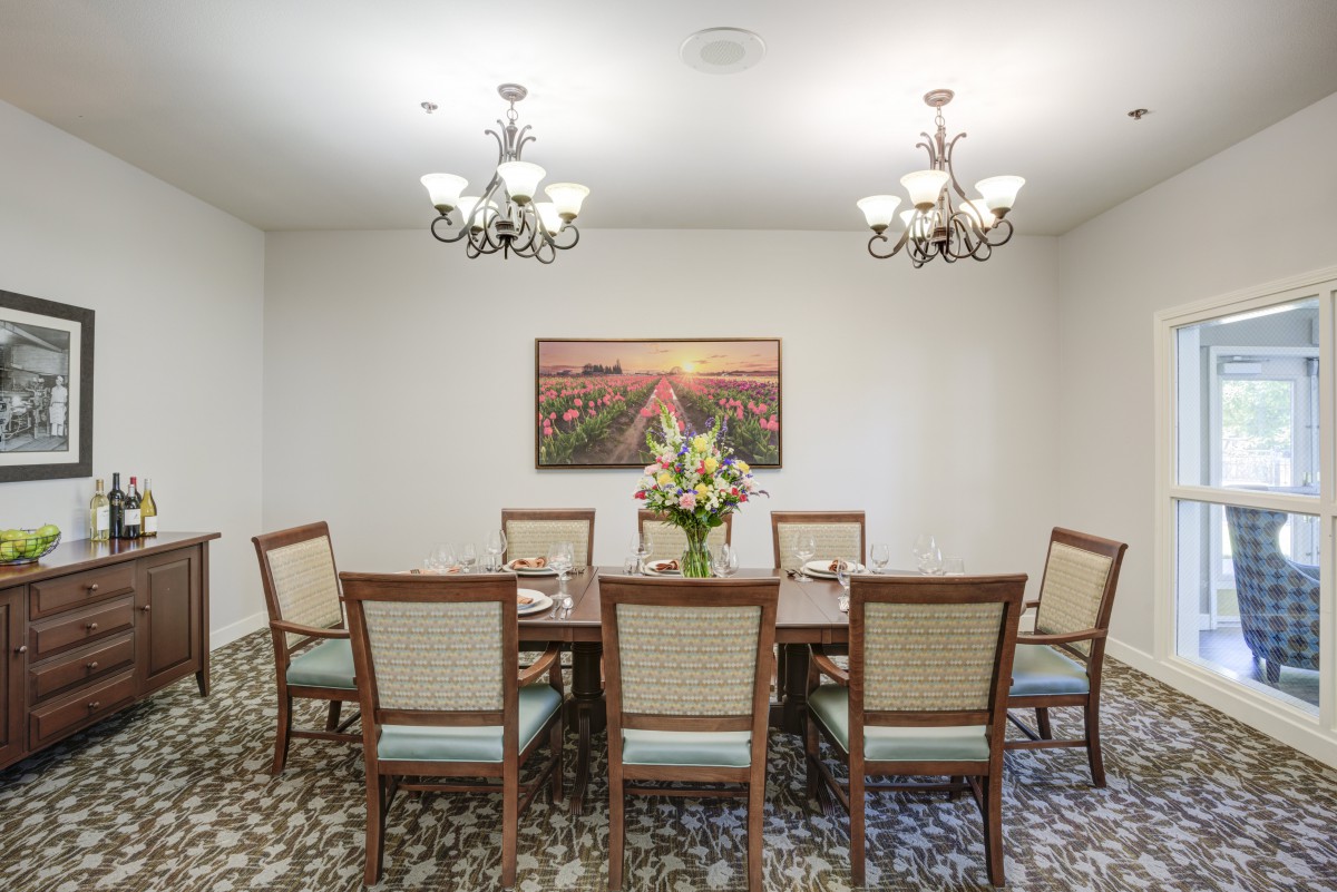 A well-lit dining room with a large table, chairs, chandeliers, and floral centerpiece beneath a scenic wall art.