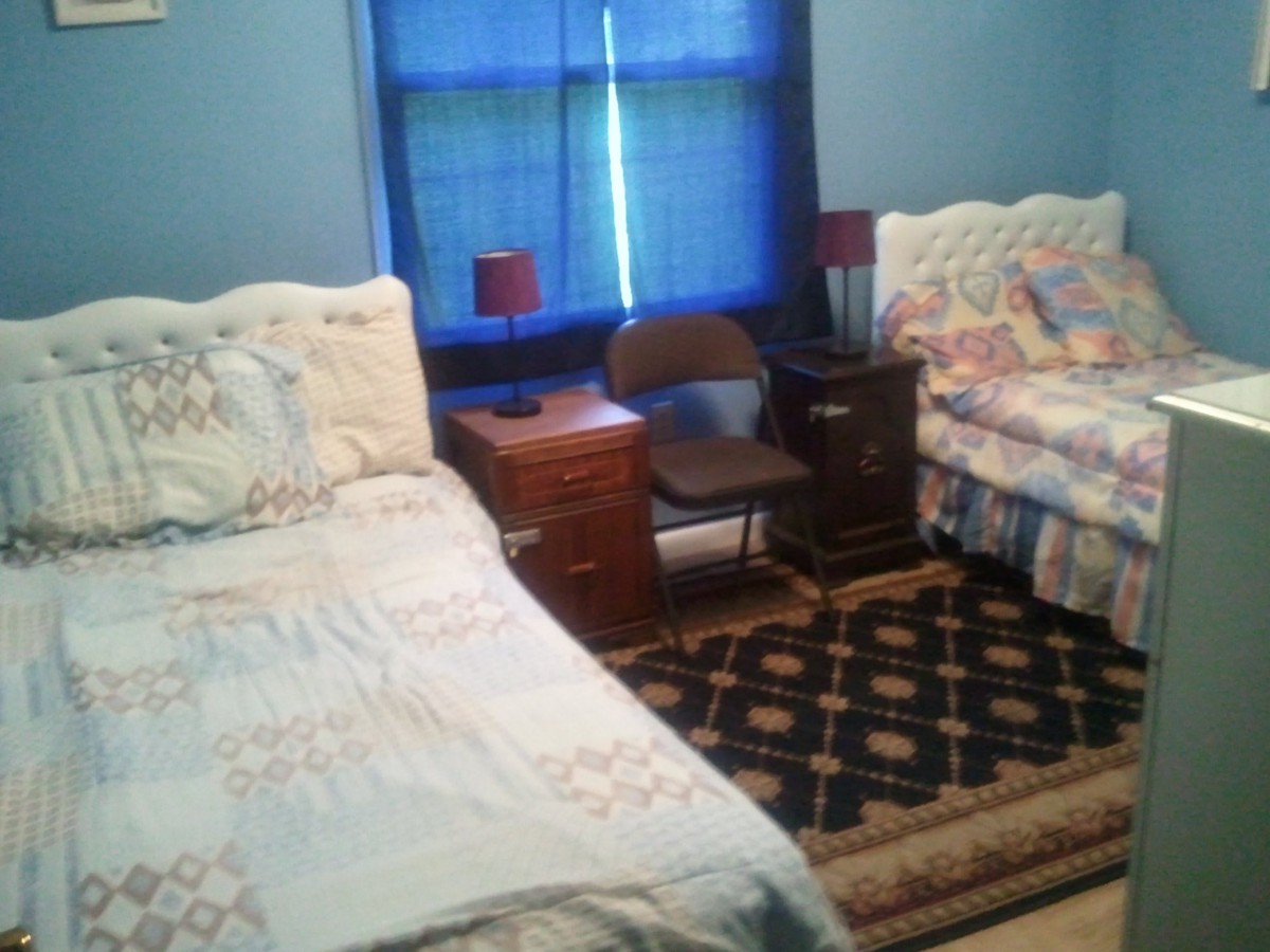A cozy bedroom featuring two beds with floral bedding, a nightstand with red lamps, a chair, and a patterned rug.