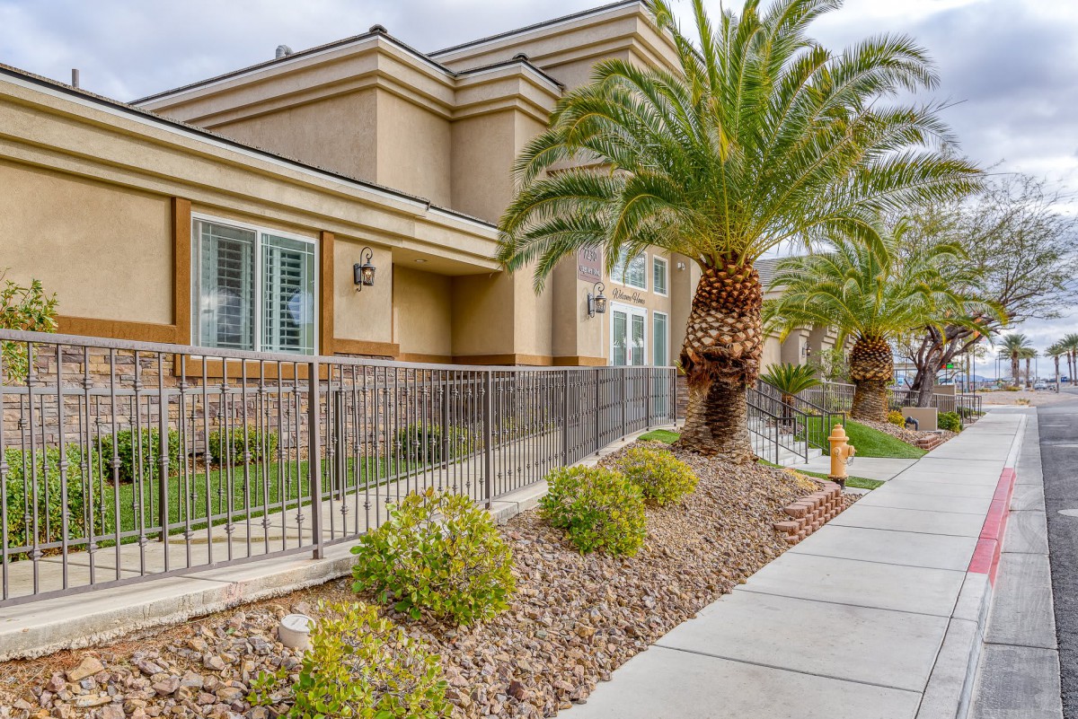 A modern residential building with landscaping features palm trees and desert plants along a fenced sidewalk.