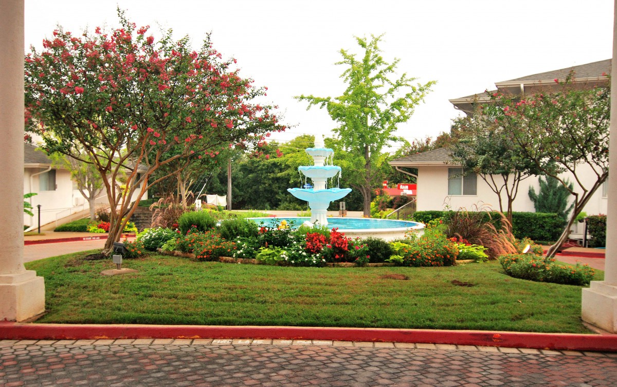 A picturesque garden featuring a multi-tiered fountain surrounded by greenery and vibrant flowers, with a house in the background.