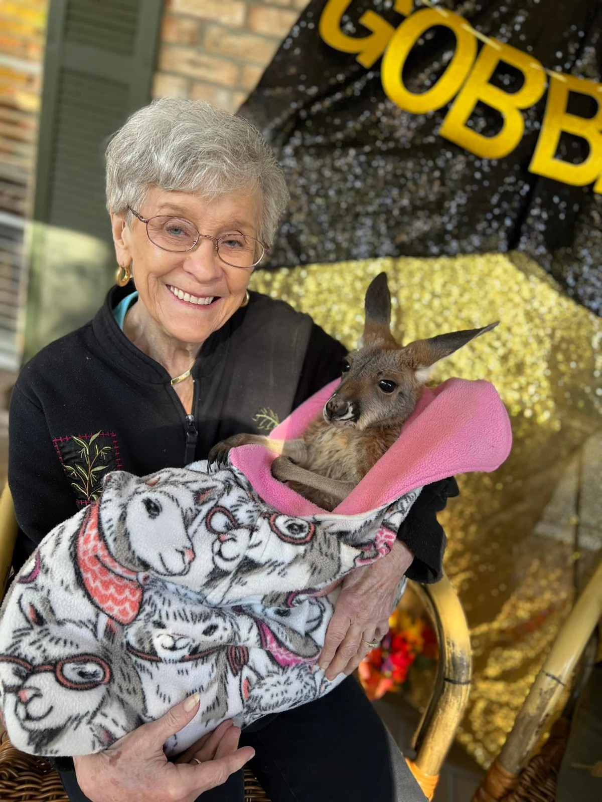 A woman smiling while holding a kangaroo wrapped in a blanket indoors.